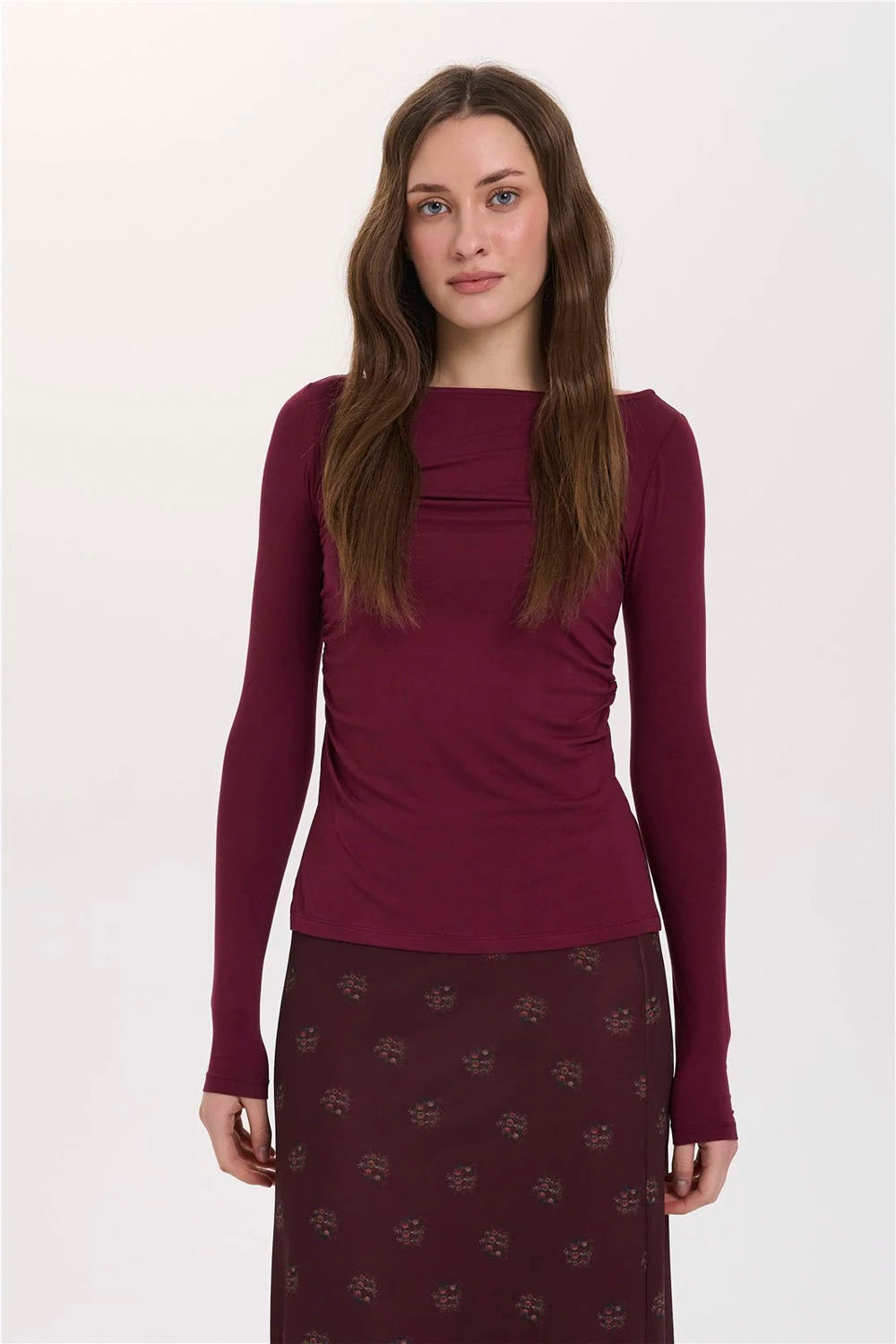 A woman modeling a long-sleeve knit top in deep burgundy with a gathered neckline, paired with a matching patterned skirt, against a plain light gray background.