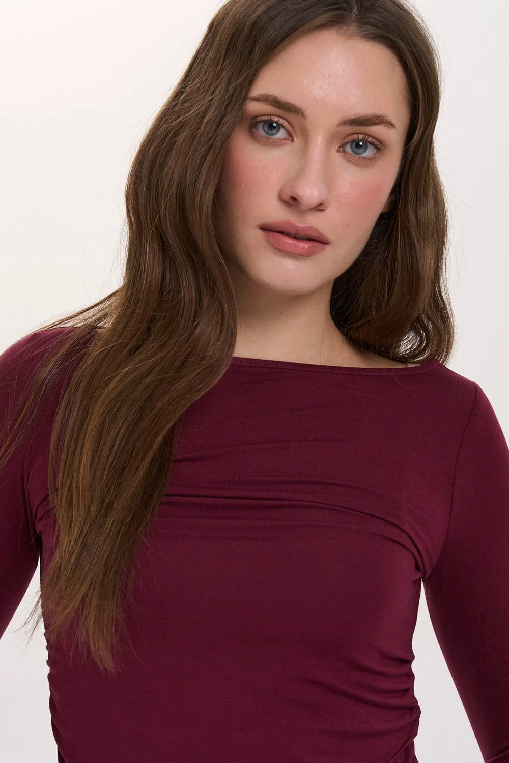 A close-up shot of a woman wearing a long-sleeve boat-neck top in a deep burgundy or plum color, set against a plain light background.