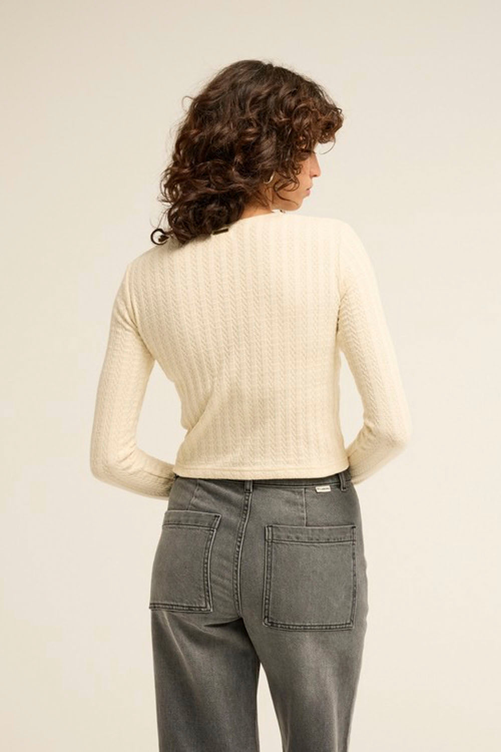 A woman shown from the back wearing a cream-colored textured knit long-sleeve top and gray jeans, set against a plain off-white studio background.