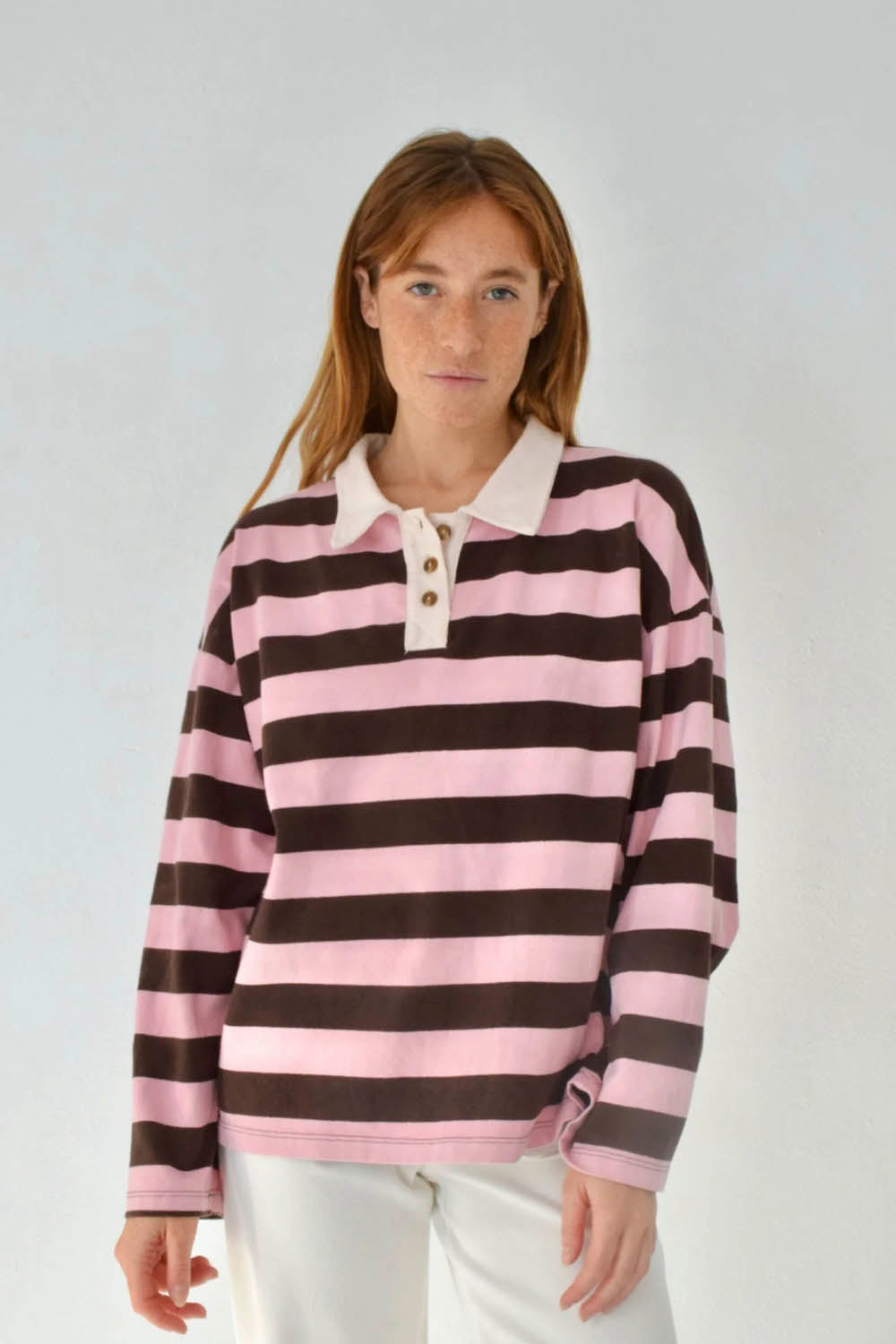 A woman wearing a pink and dark brown horizontally striped long-sleeve polo shirt with a cream collar and buttons, paired with white pants, against a light gray studio background.