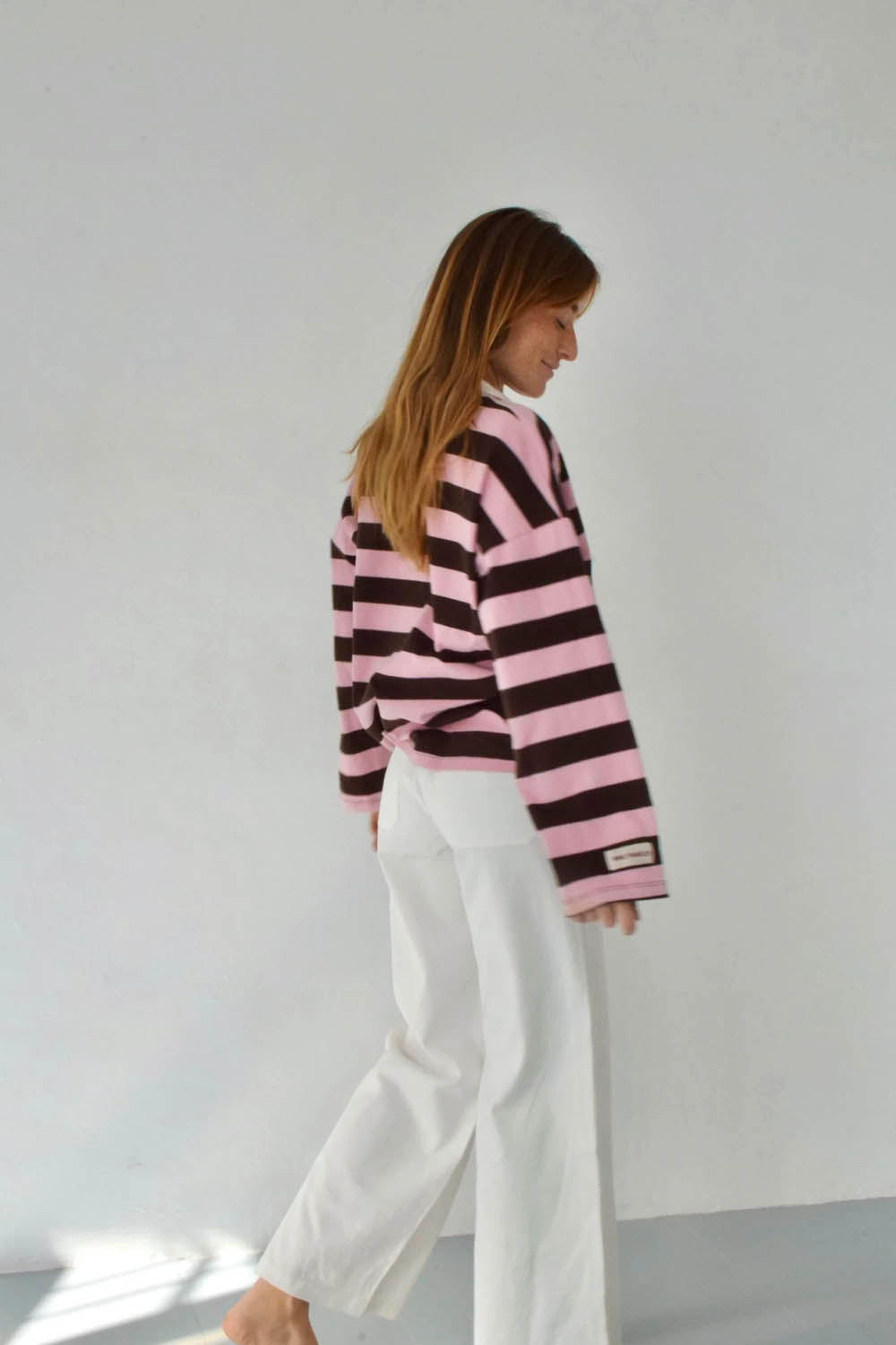 A woman wearing a pink and dark brown striped long-sleeve knit top with white wide-leg pants, photographed from the back in a minimalist studio setting.