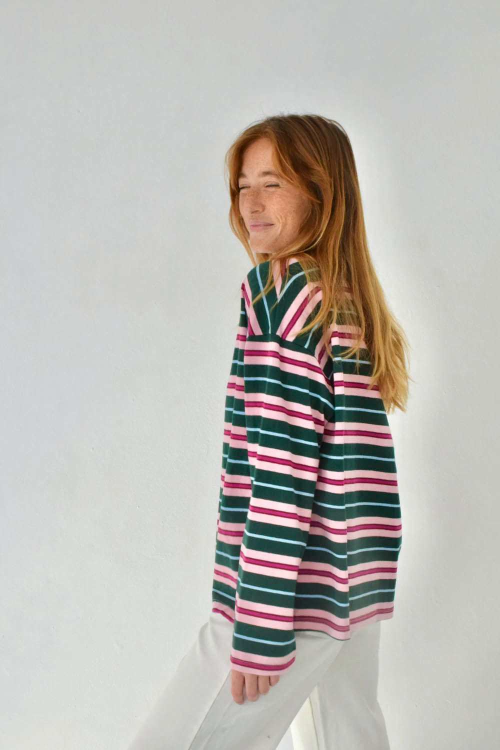 A woman with long light brown hair is shown from the side, wearing a striped knit top with horizontal bands of dark green, pink, and white, paired with white pants, against a plain light gray background.