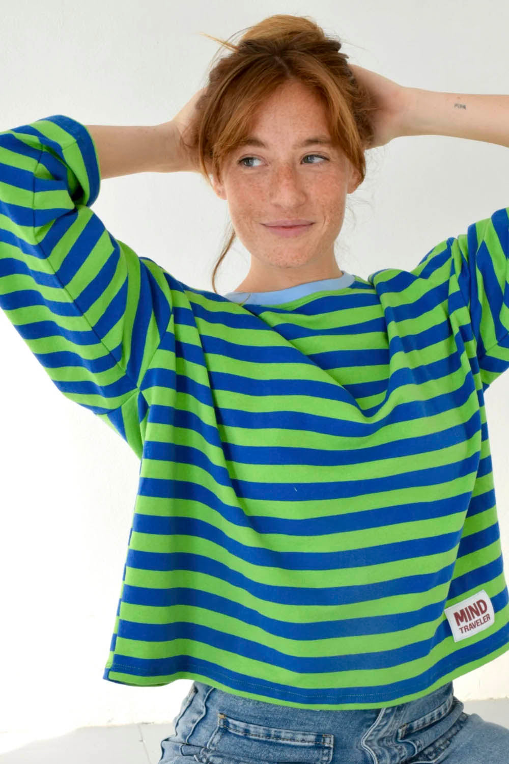 A woman wearing a long-sleeve knit top with horizontal lime green and blue stripes, paired with light-wash blue jeans, standing against a plain light gray wall.