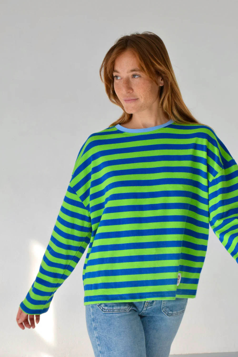 A woman wearing a long-sleeve knit top with horizontal lime green and blue stripes, paired with light-wash blue jeans, standing against a plain light gray wall.