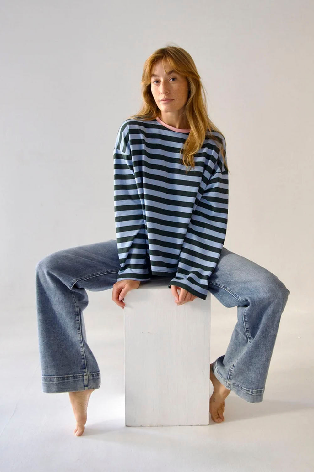 A woman with long light brown hair sits barefoot on a white block, wearing a navy and gray striped long-sleeve shirt and wide-leg light blue jeans against a plain light gray studio background.