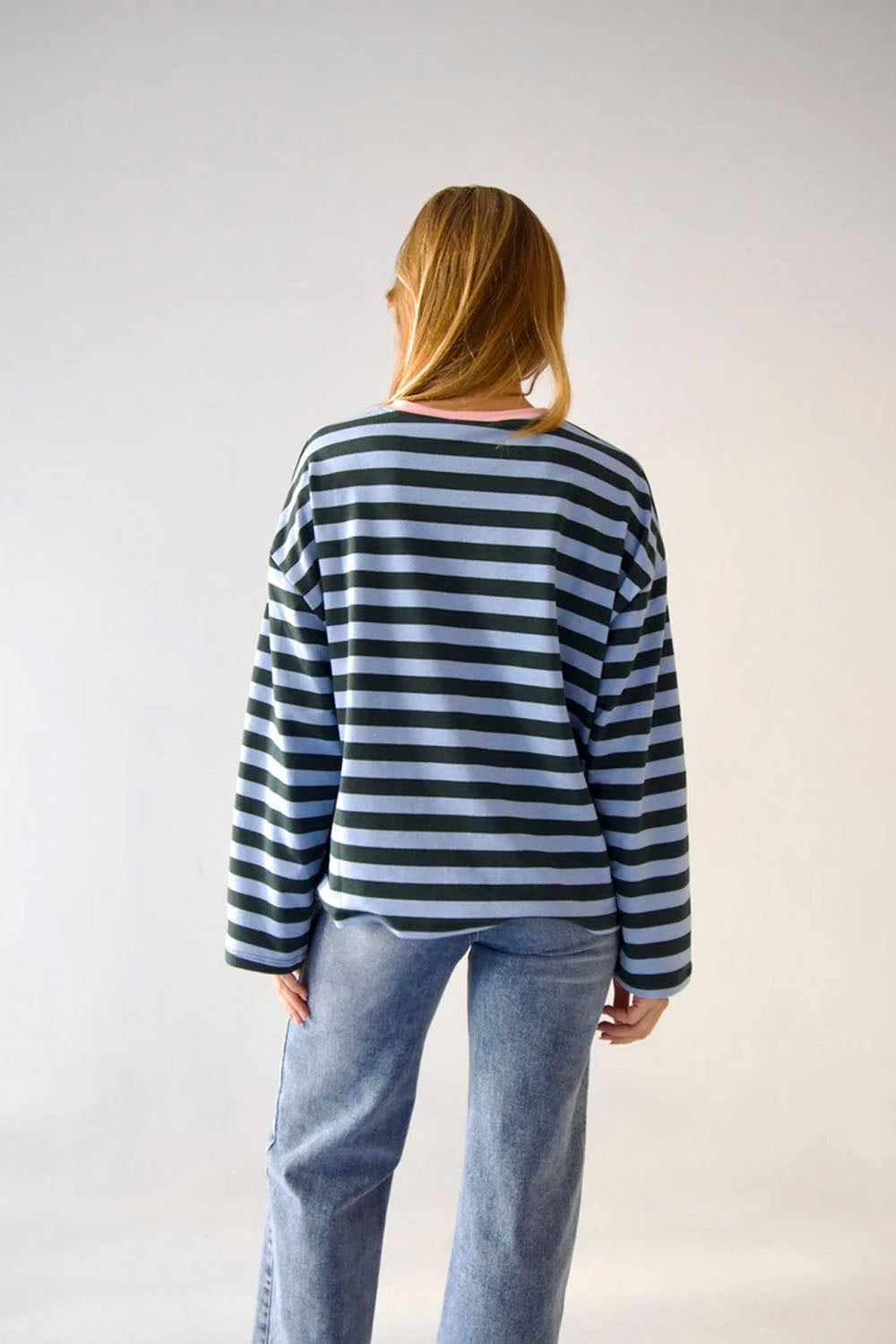 A woman viewed from behind wearing a navy and light blue horizontally striped long-sleeve top with light-wash blue jeans, standing against a plain light gray studio background.