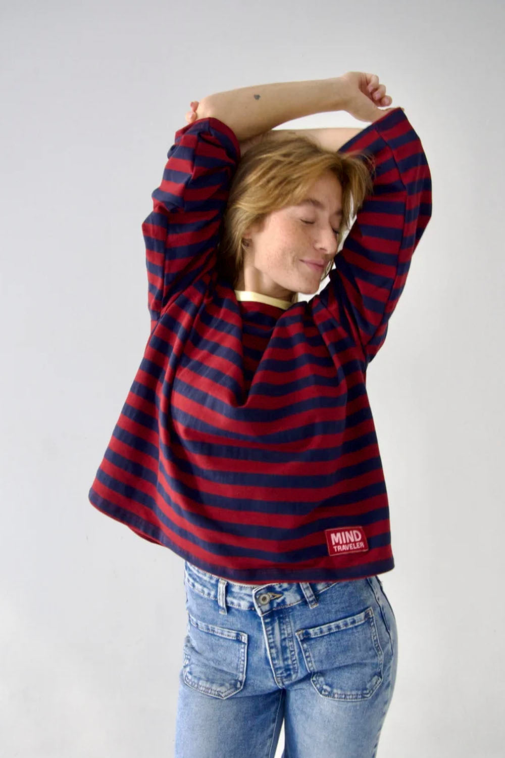 A woman wearing a red and navy blue horizontally striped long-sleeve sweatshirt with a small red 'MIND' brand patch on the bottom hem, paired with blue jeans, posing with her arms raised against a plain light gray background.
