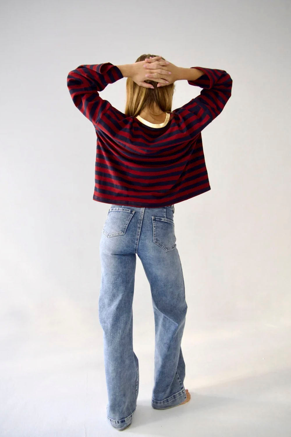 A person standing with their back to the camera, wearing a red and black horizontally striped long-sleeve crop top and light blue jeans, against a plain light gray studio background.