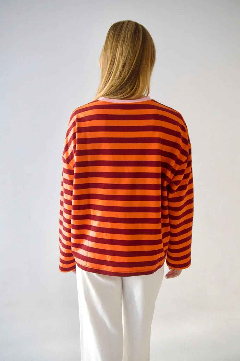 A woman viewed from behind wearing a long-sleeve knit top with horizontal orange and dark red stripes, paired with white pants, against a plain light gray studio background.