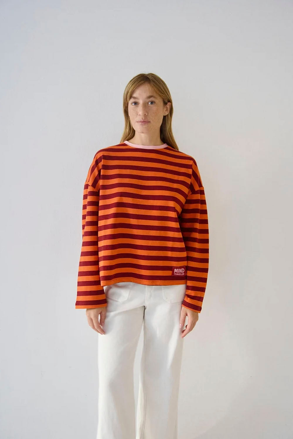 A woman modeling a long-sleeve knit top with horizontal orange and red stripes, paired with white pants, against a plain light gray background.