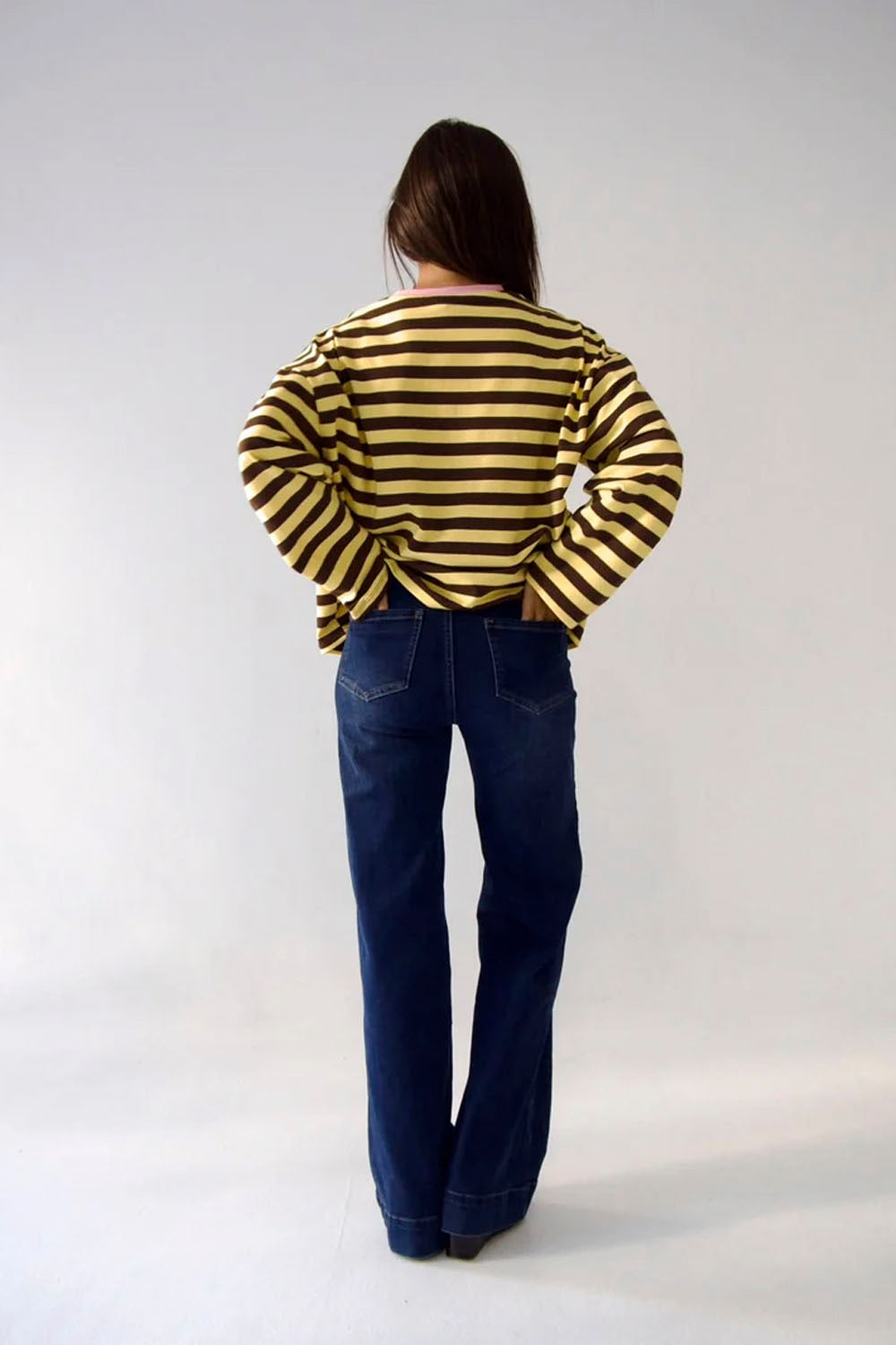 A person standing with their back to the camera, wearing a yellow and black horizontally striped long-sleeve top and dark blue jeans, against a plain light gray studio background.