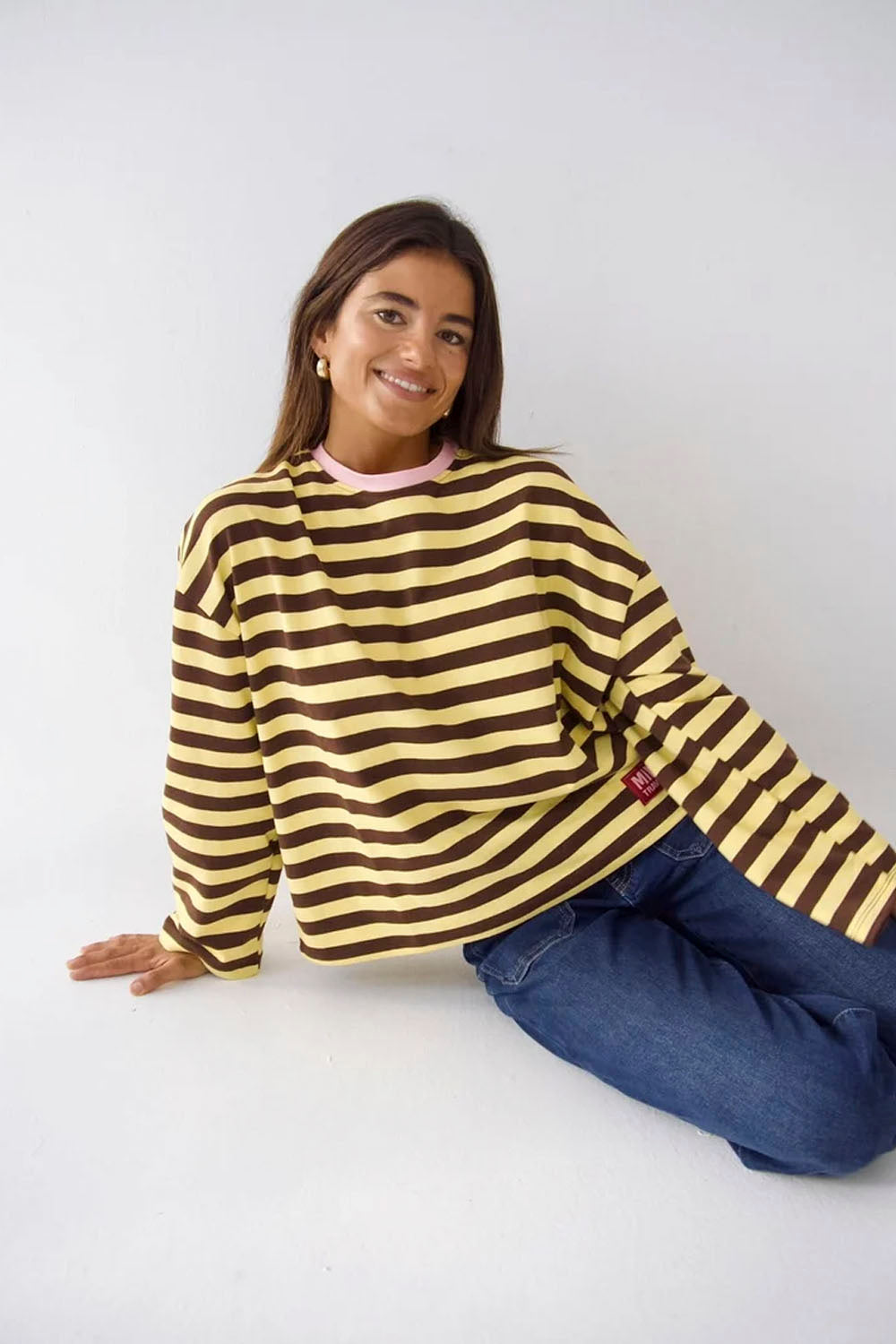 A woman wearing a brown and yellow striped long-sleeve sweatshirt and blue jeans, sitting on a white studio floor against a white background.