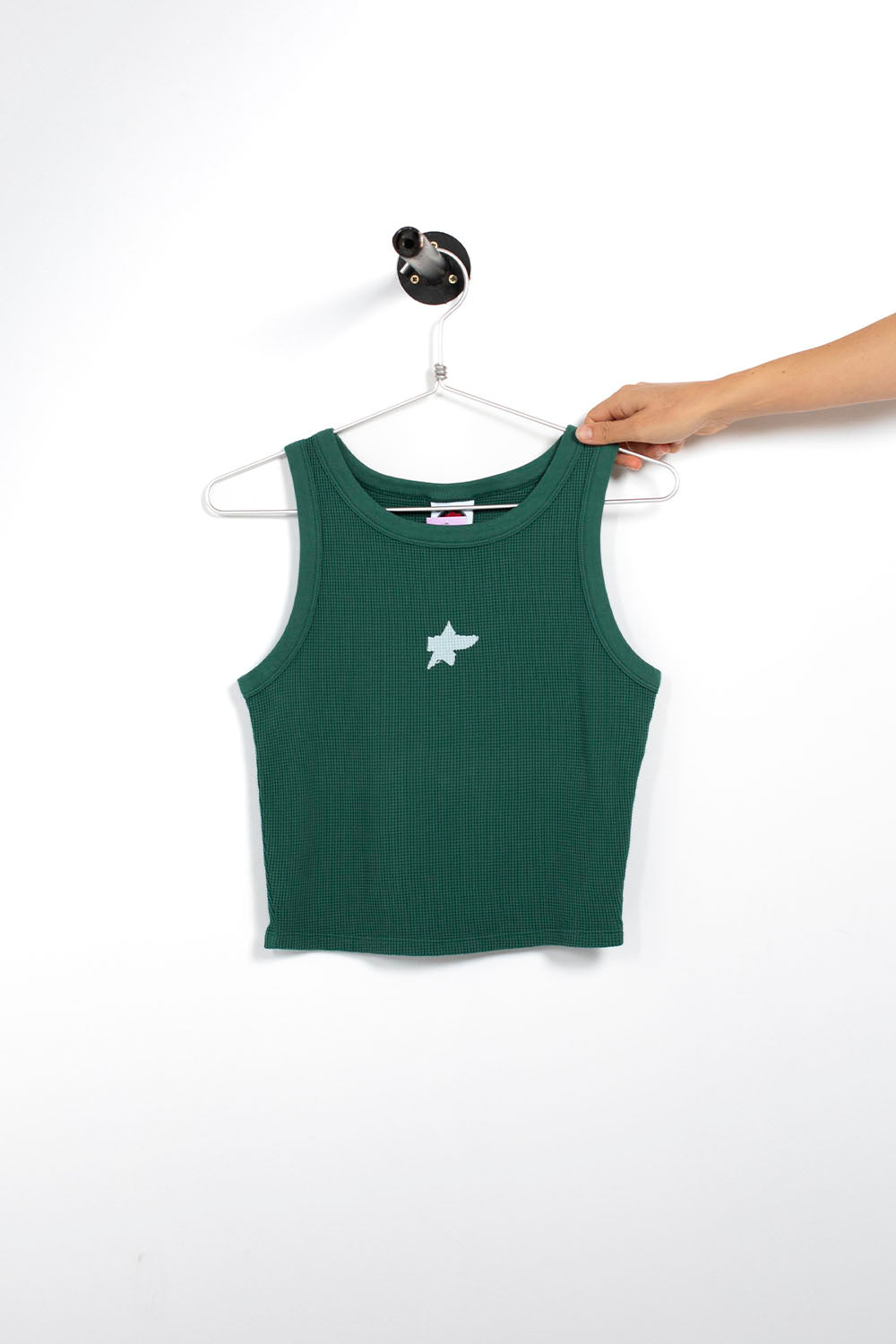 A dark green ribbed sleeveless crop top with a small white star graphic on the chest, hanging on a white hanger held by a person's hand against a plain white wall.