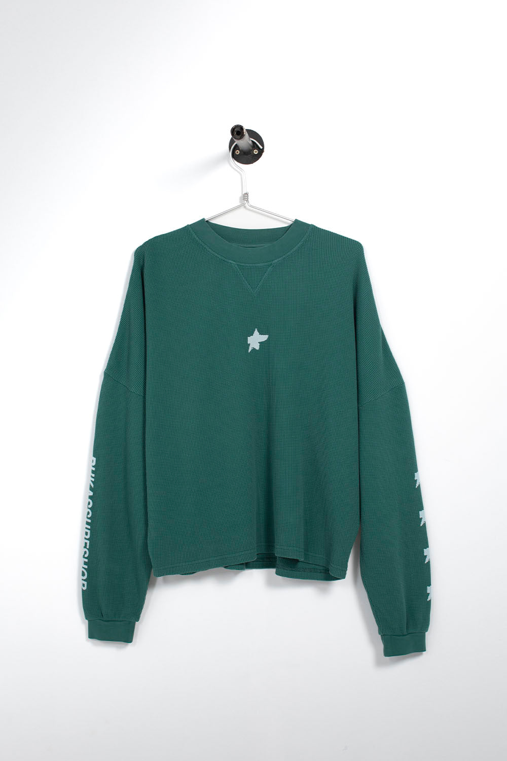 A dark green long-sleeve sweatshirt with a small white star graphic on the chest and white text/graphic details on the sleeves, hanging on a white hanger against a white wall.