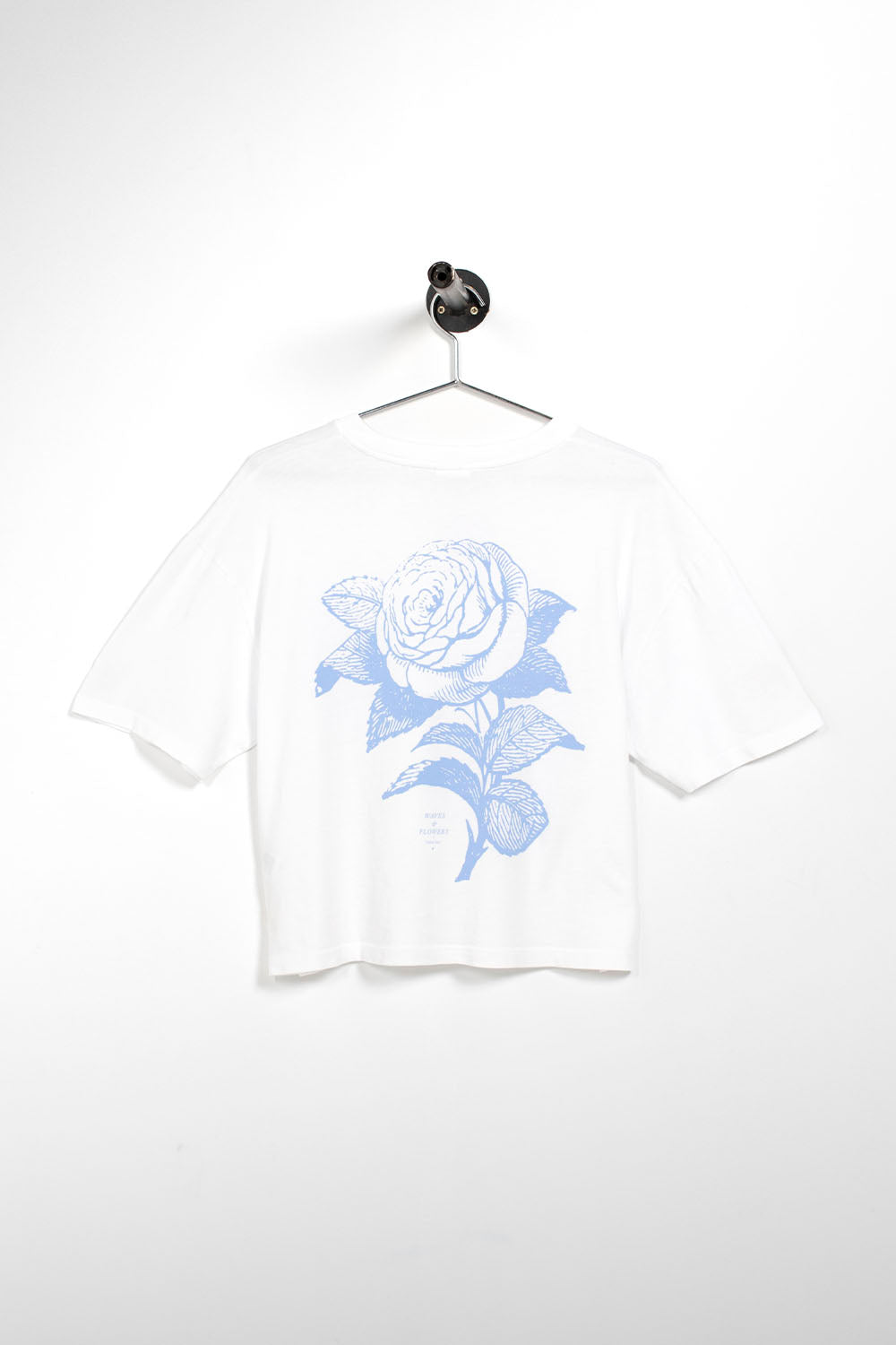 A white short-sleeve crop top t-shirt hanging on a metal hanger against a white wall. The shirt features a large light blue line-art graphic of a rose on the front.