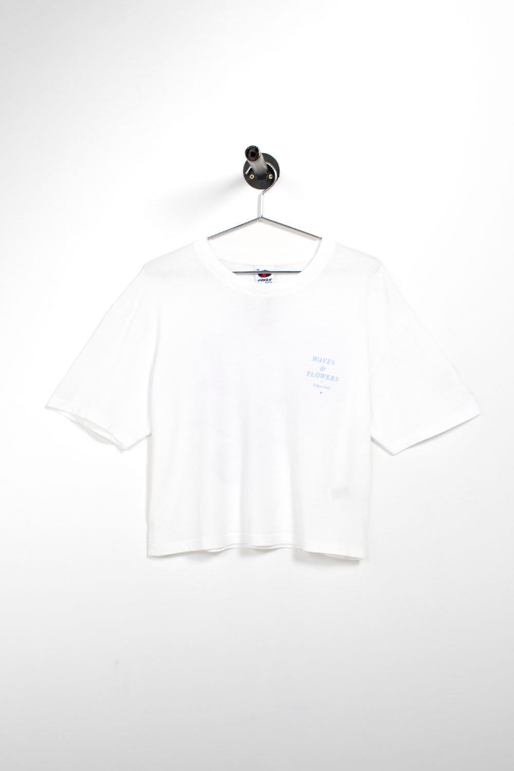 A white short-sleeve crop top t-shirt with a small light blue graphic on the left chest, hanging on a metal hanger against a white wall.