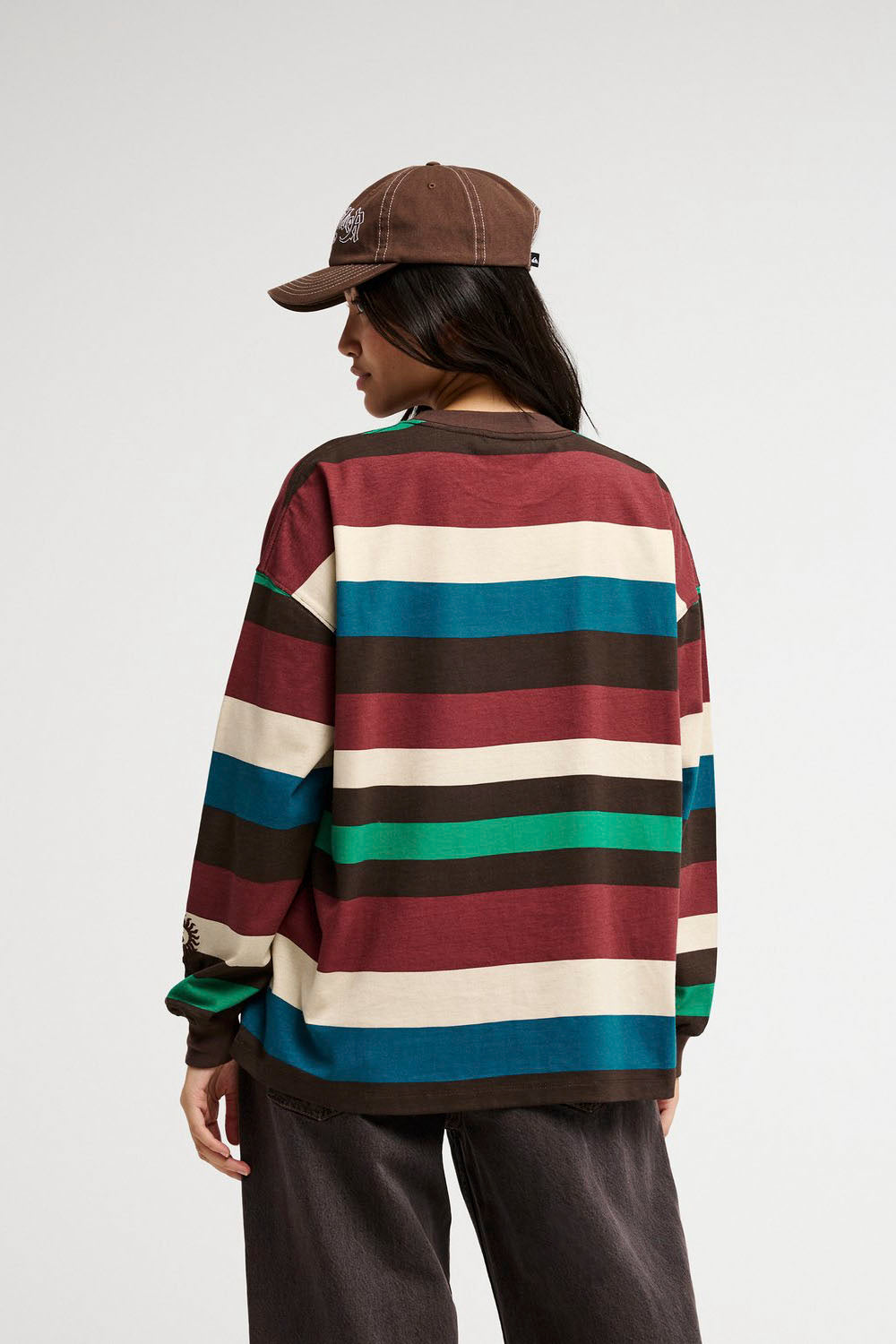 A model from the back wearing a long-sleeve crewneck shirt with horizontal stripes in burgundy, cream, dark brown, teal, and green, paired with dark brown pants and a brown baseball cap, against a light gray studio background.
