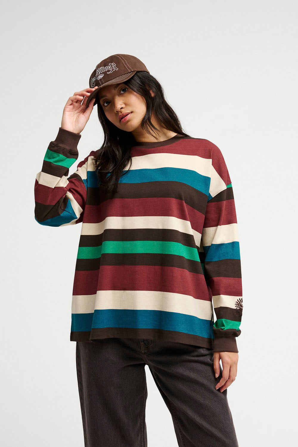 A woman wearing a brown baseball cap, a long-sleeve crewneck shirt with horizontal stripes in brown, burgundy, teal, black, and cream, and dark brown pants, posing against a light gray background.