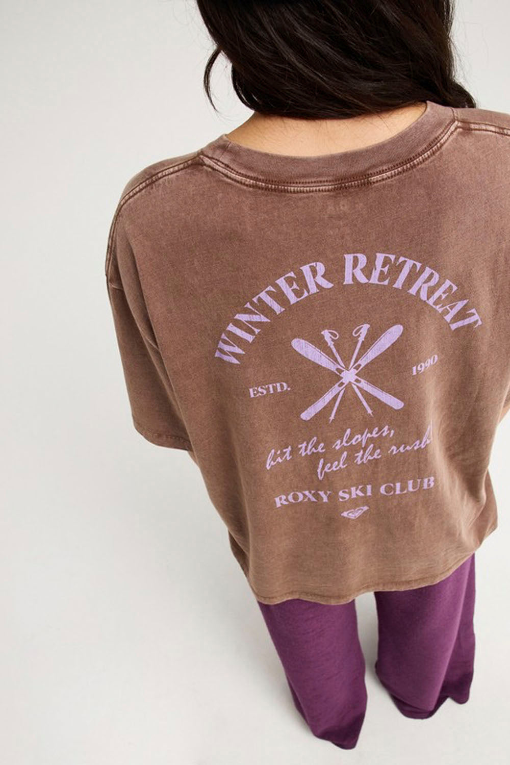 A person shown from the back wearing a brown acid-wash graphic t-shirt with purple text and graphics reading 'WINTER RETREAT' and 'ROXY SKI CLUB', paired with purple pants, against a plain light gray background.