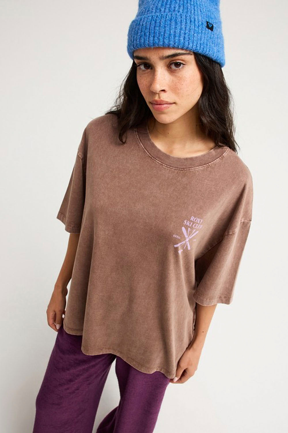 A woman wearing a brown mineral-wash oversized t-shirt with a small white graphic on the left chest, paired with purple pants and a blue knit beanie, photographed against a plain white studio background.