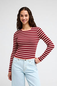 A woman modeling a long-sleeve ribbed knit top with horizontal red and white stripes, paired with light blue jeans, against a plain light gray studio background.