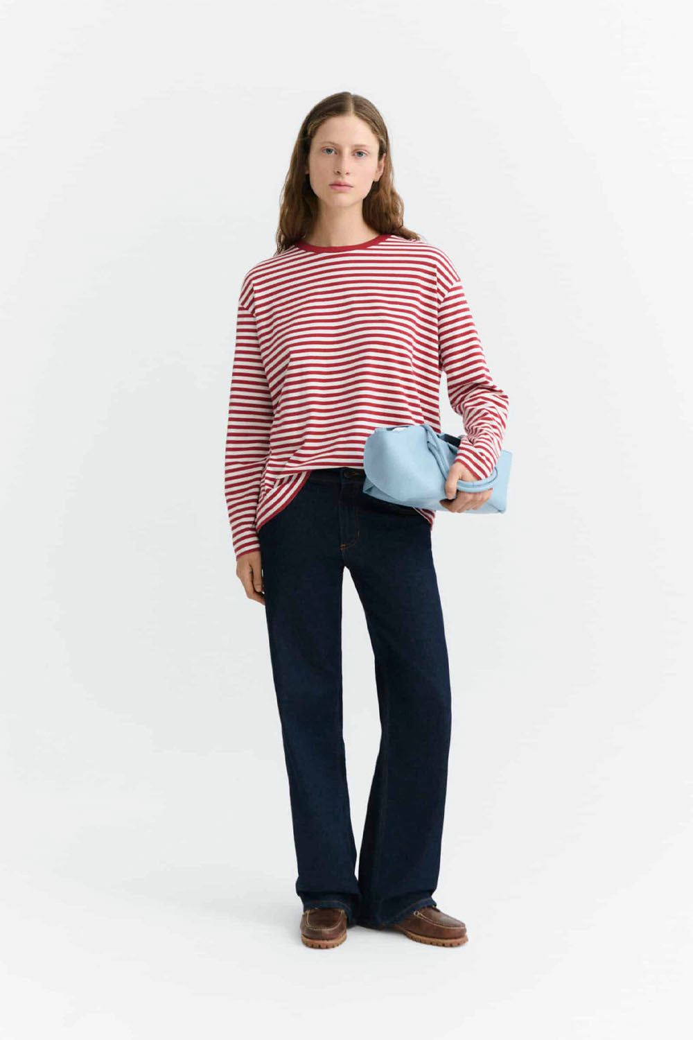 A woman modeling a red and white horizontally striped long-sleeve top, dark blue jeans, and brown shoes, holding a light blue clutch bag against a plain white studio background.