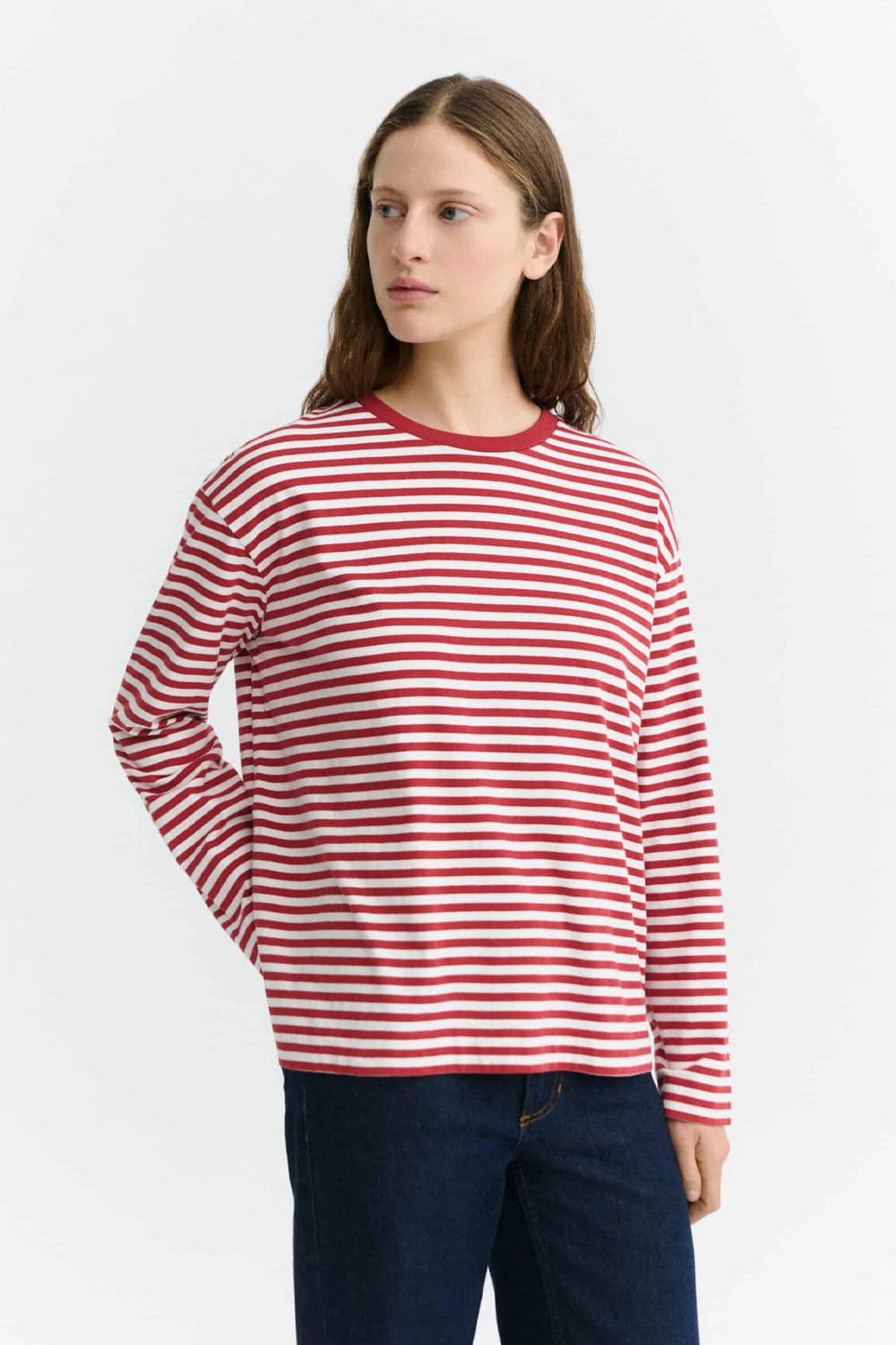 A woman wearing a red and white horizontally striped long-sleeve crewneck shirt with dark blue jeans, standing against a plain white studio background.