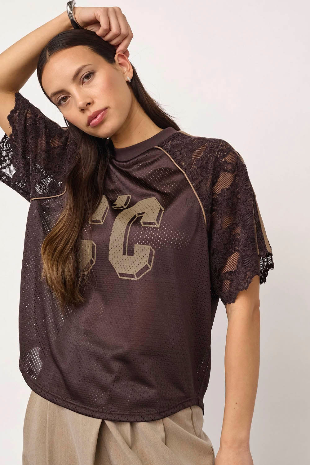 A woman wearing a dark brown mesh short-sleeve top with lace sleeves and a large beige number 22 graphic on the front, paired with beige pants, against a plain light background.