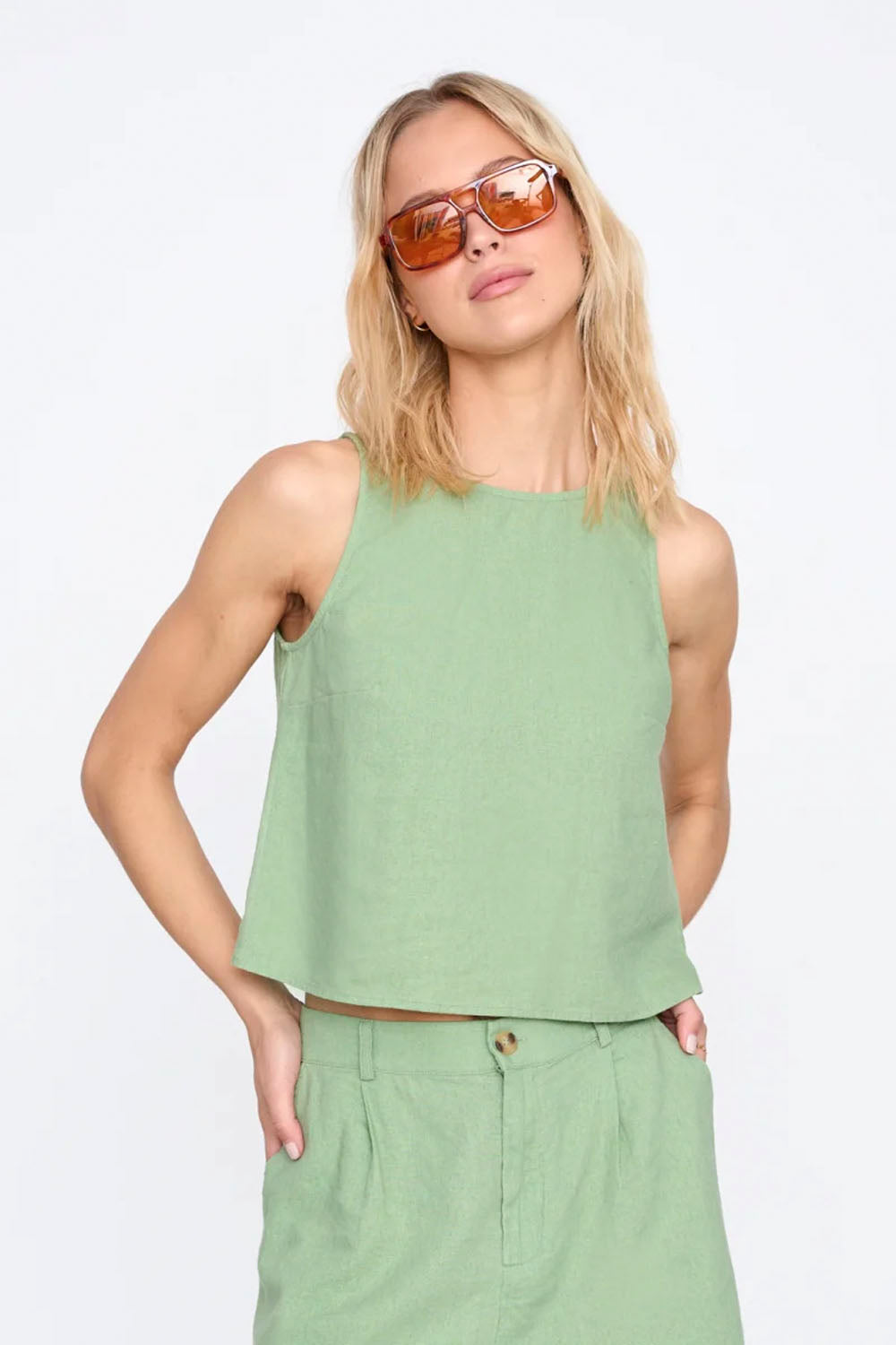 Woman wearing a light green sleeveless top and pants with sunglasses on a white background