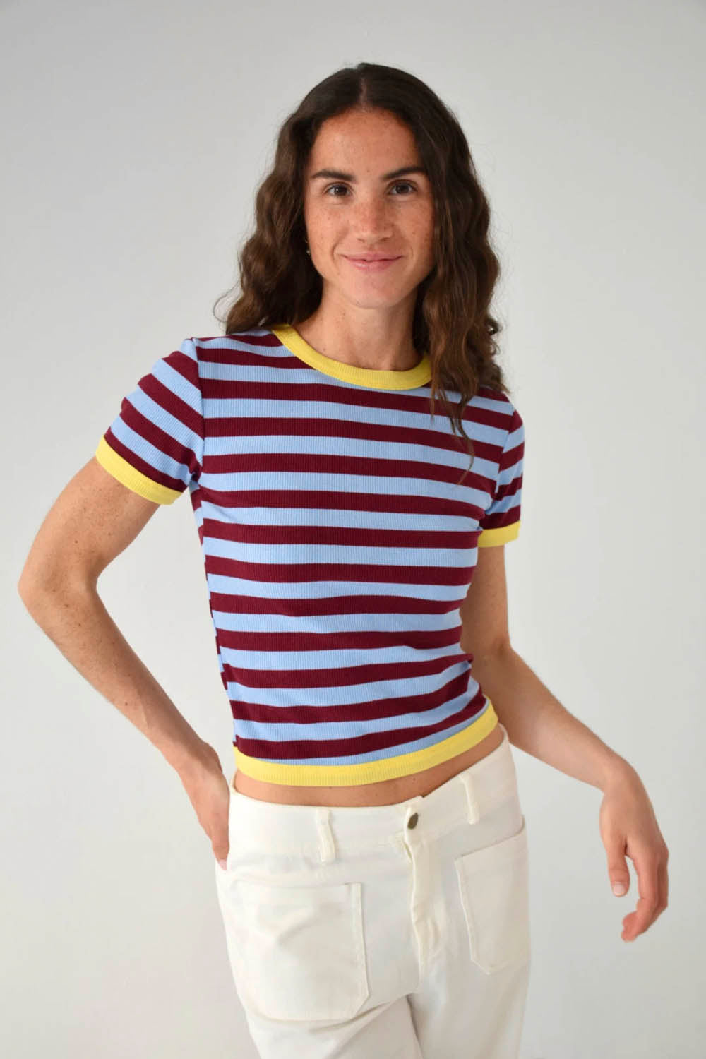 A woman modeling a short-sleeve ringer t-shirt with horizontal burgundy and light blue stripes, featuring yellow trim on the collar and sleeve cuffs, paired with white trousers against a plain light gray background.