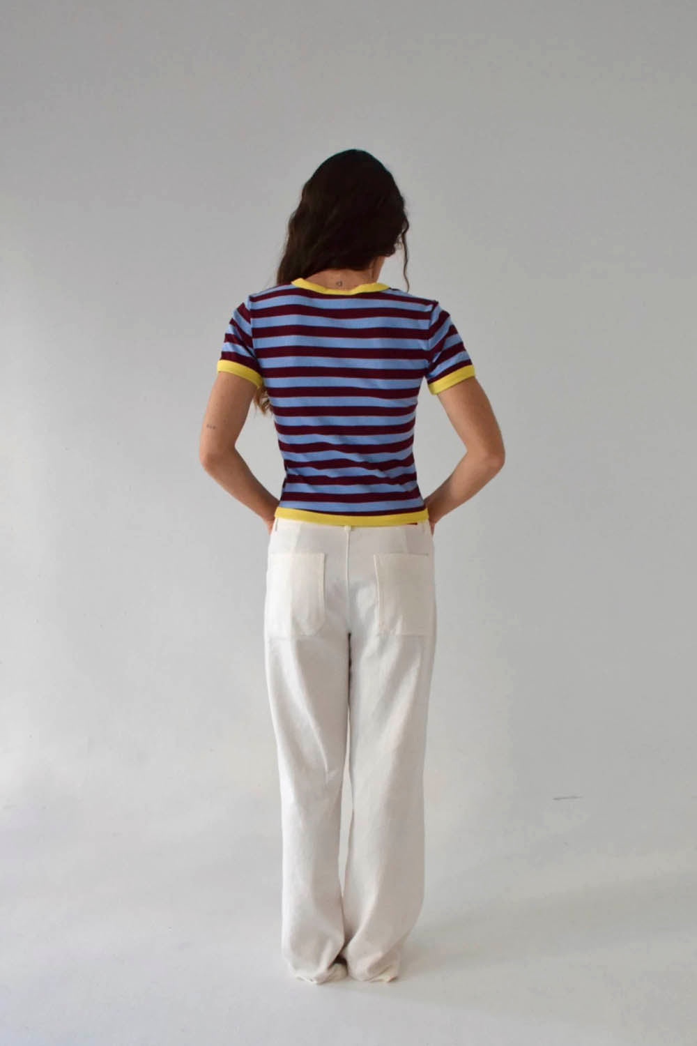 A woman modeling a short-sleeve ringer t-shirt with horizontal burgundy and light blue stripes, featuring yellow trim on the collar and sleeve cuffs, paired with white trousers against a plain light gray background.