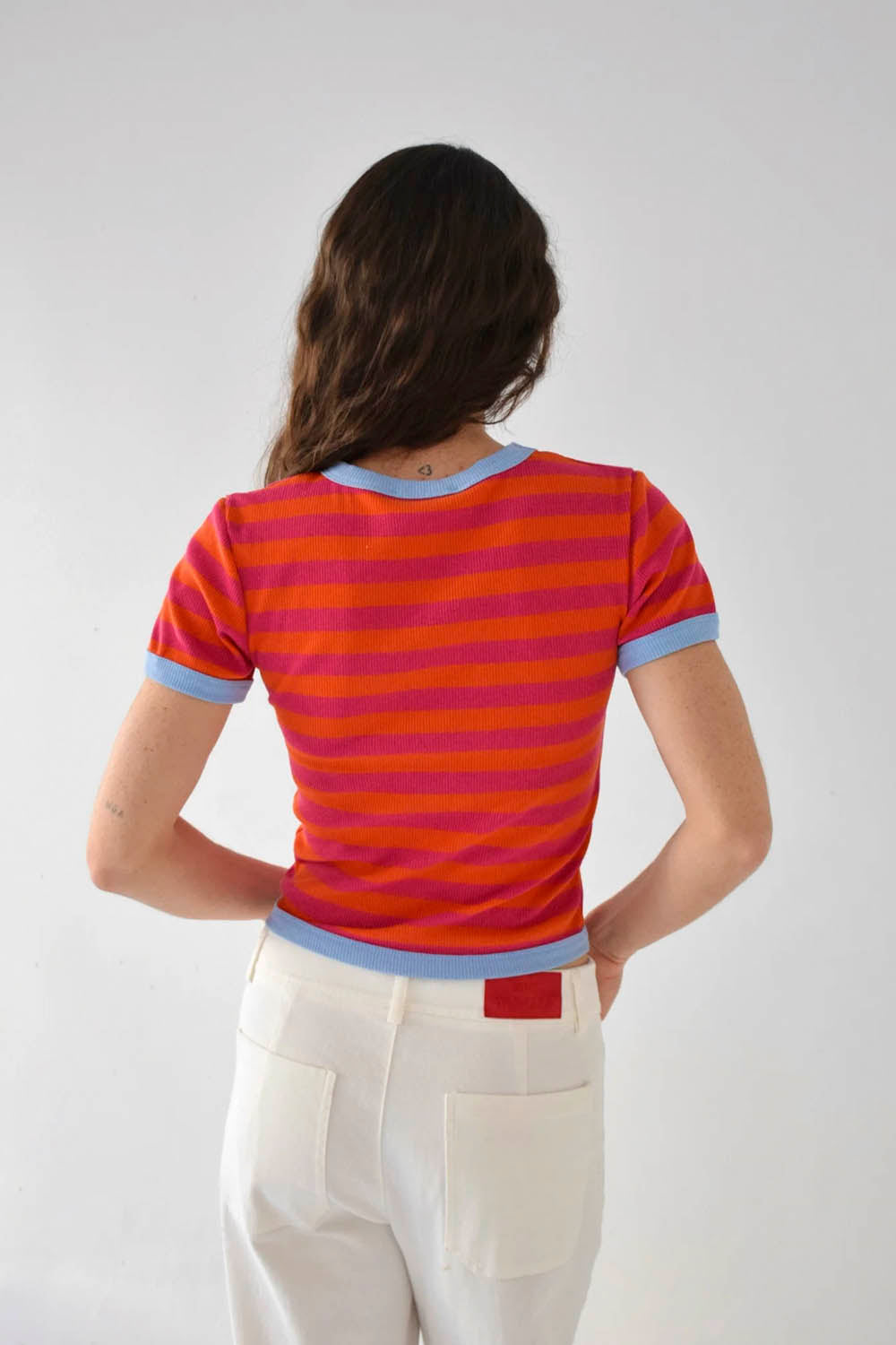 A woman wearing a short-sleeve crewneck t-shirt with horizontal orange and pink stripes and light blue ribbed trim, paired with white pants, against a light gray studio background.