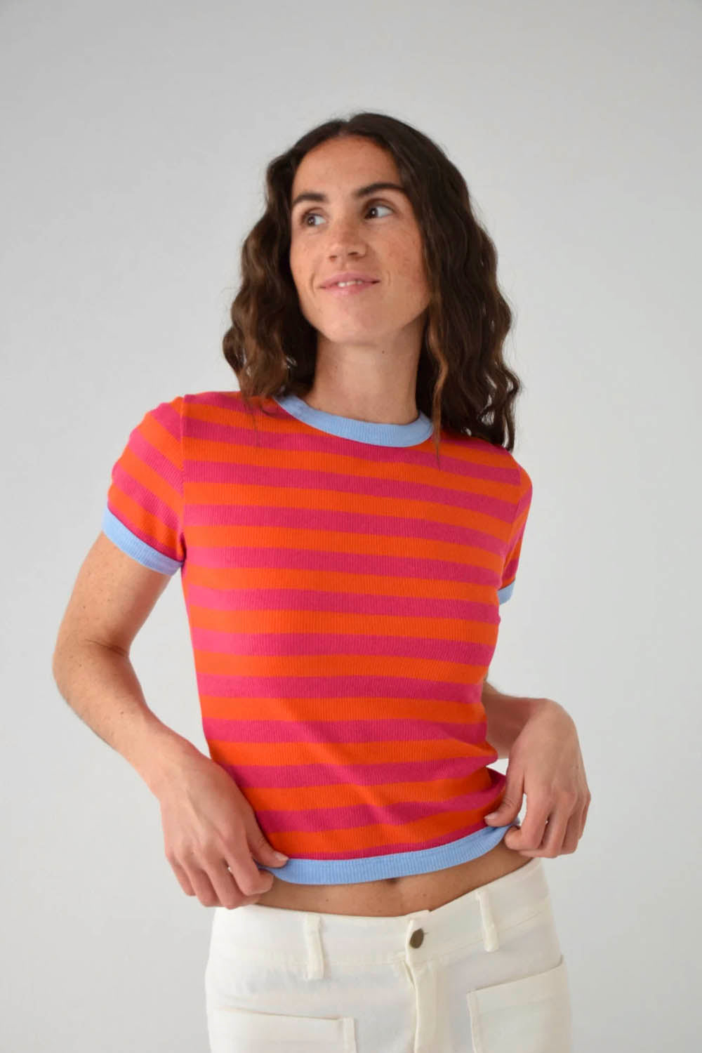 A woman wearing a short-sleeve crewneck t-shirt with horizontal orange and pink stripes and light blue ribbed trim, paired with white pants, against a light gray studio background.