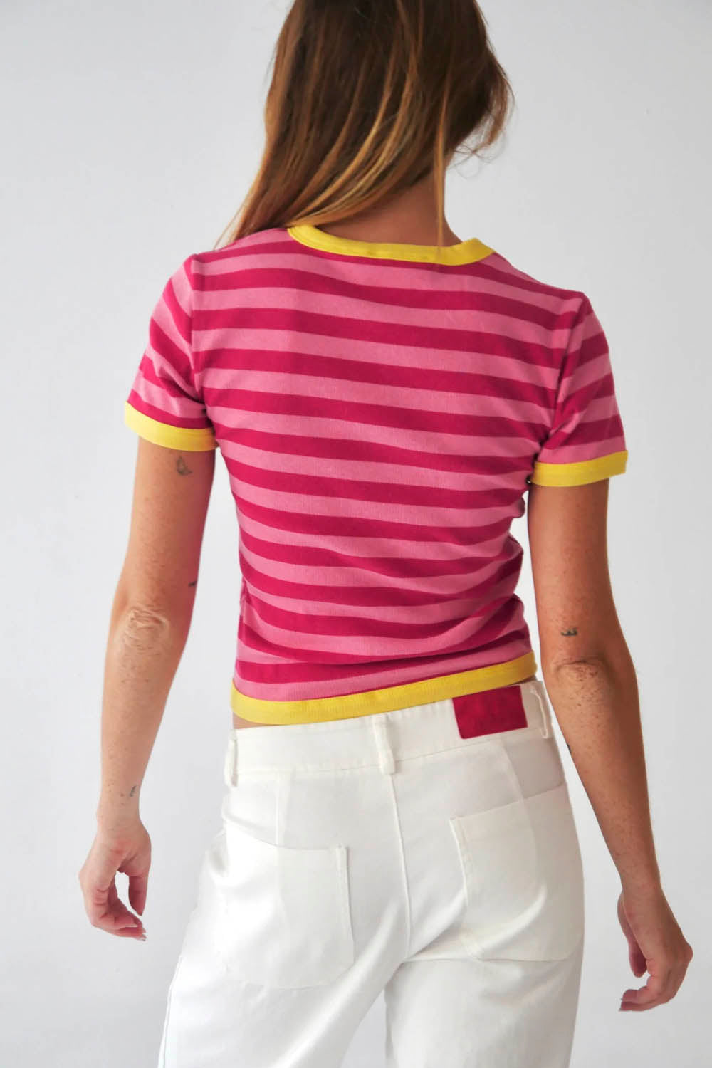 A woman wearing a pink and fuchsia striped ringer t-shirt with yellow trim, paired with white pants, standing against a plain light gray background.