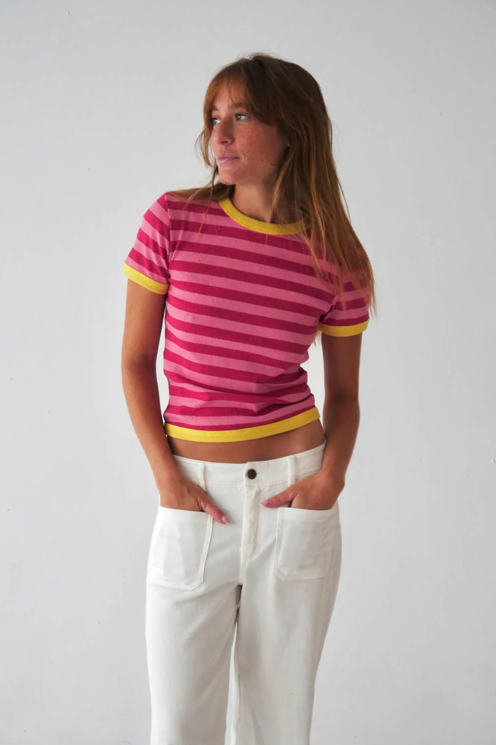 A woman wearing a pink and fuchsia striped ringer t-shirt with yellow trim, paired with white pants, standing against a plain light gray background.