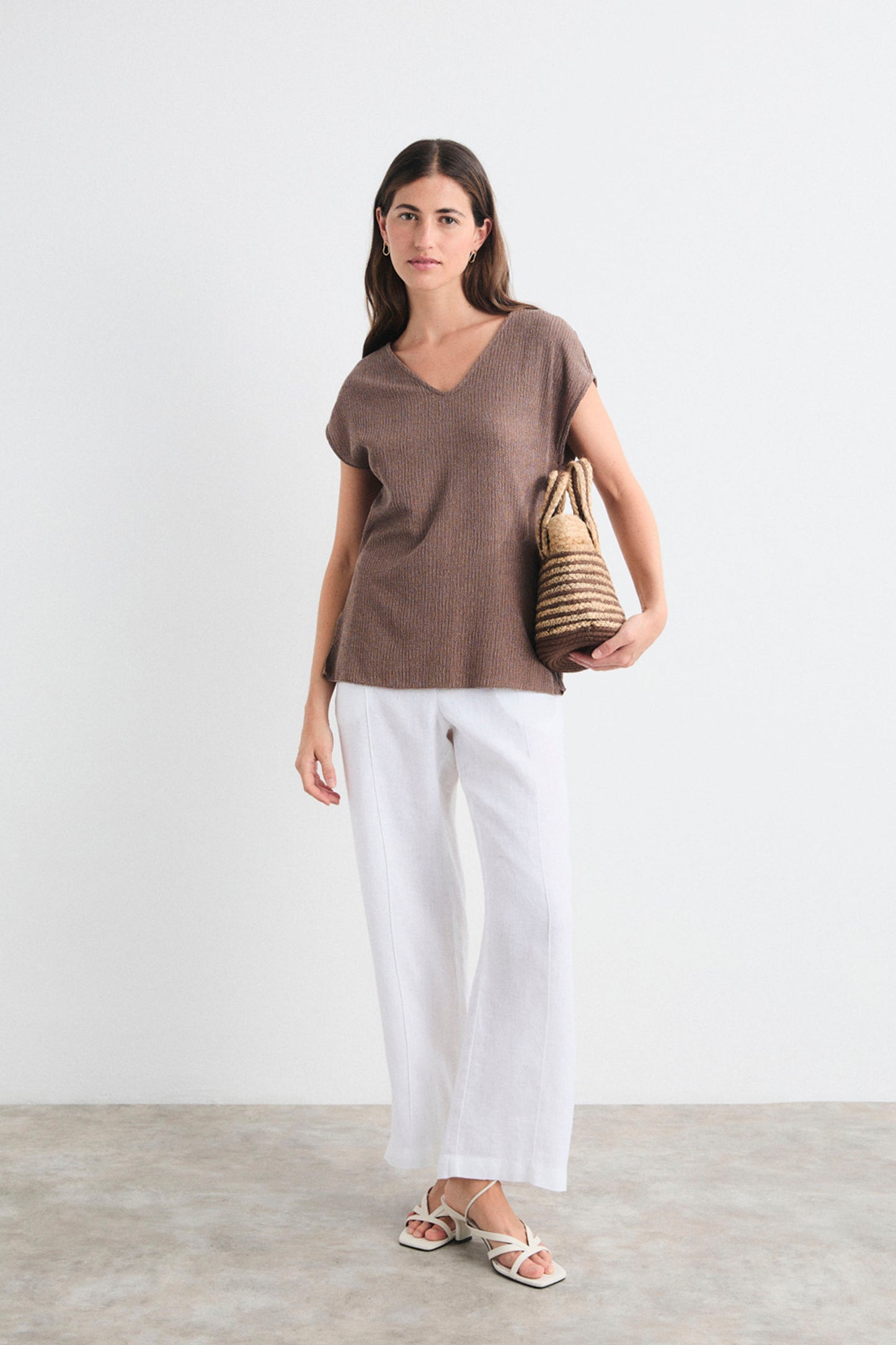Woman wearing a brown top and white pants holding a woven bag on a plain background