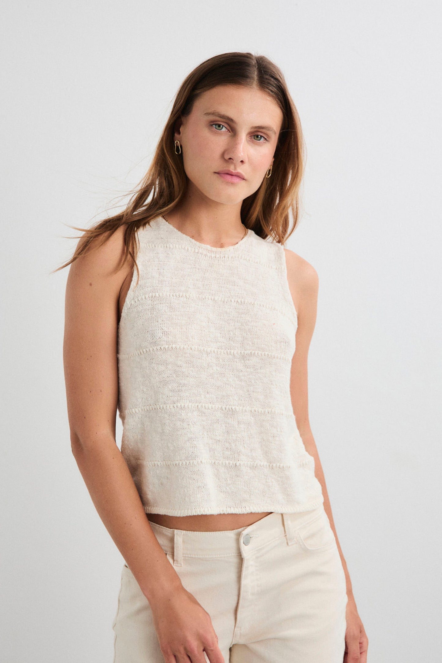 Woman wearing a sleeveless white top and beige pants against a plain background