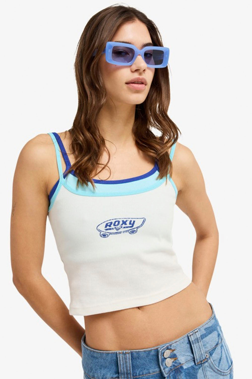 Woman wearing a white tank top with blue accents and 'Roxy' branding, paired with blue sunglasses and jeans on a white background.