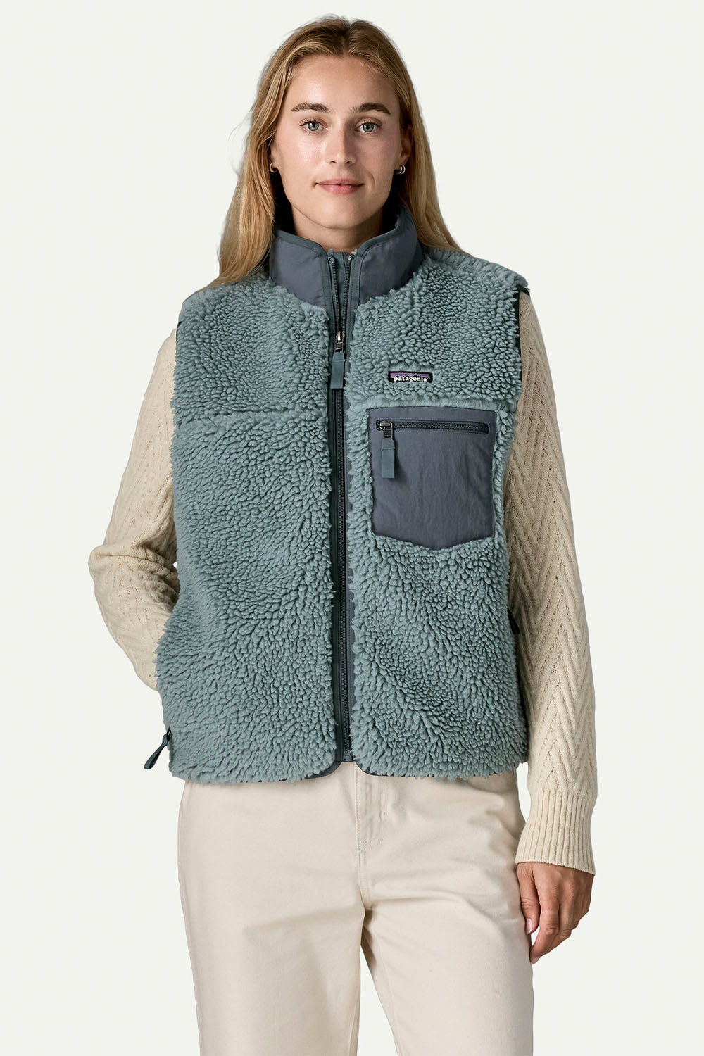 A woman modeling a Patagonia fleece vest in a dusty teal-blue color, worn over a cream cable-knit sweater and cream pants against a light gray studio background.