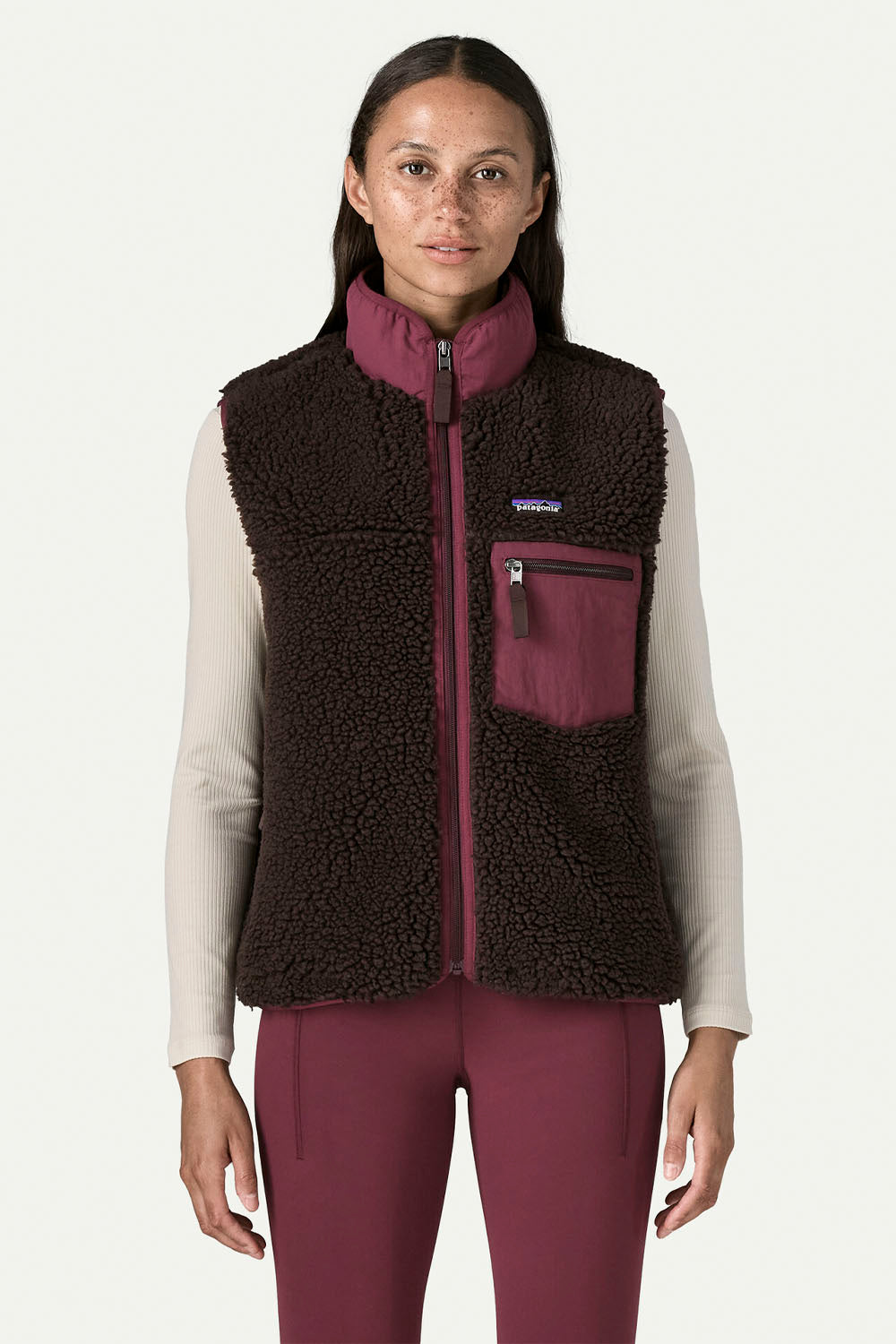 A woman modeling a Patagonia fleece vest in dark brown and burgundy, worn over a light beige long-sleeve shirt and matching burgundy pants, against a clean off-white studio background.