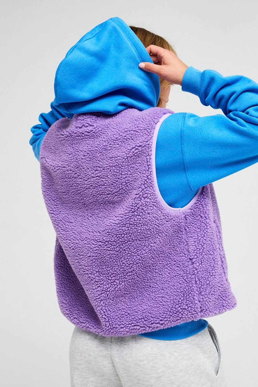 A person shown from the back wearing a bright blue hoodie layered over a lavender purple fleece vest, paired with light gray sweatpants, against a plain light gray background.
