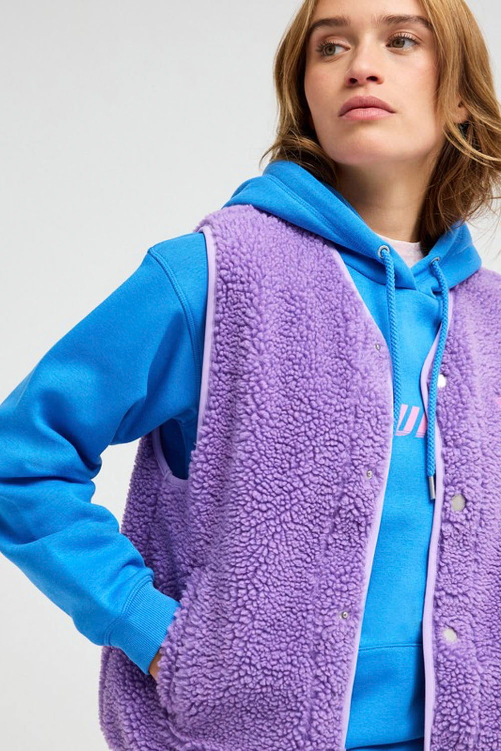 A close-up of a model wearing a lavender purple sherpa fleece zip-up vest over a bright blue hoodie, set against a plain light gray studio background.