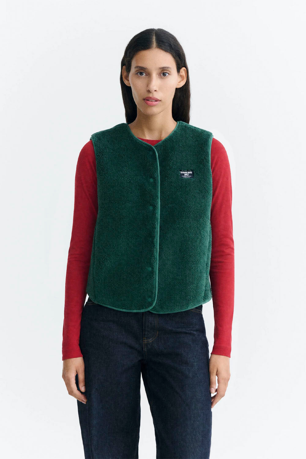 A model wearing a dark green fleece vest over a red long-sleeve shirt and dark blue jeans, photographed against a clean white studio background.