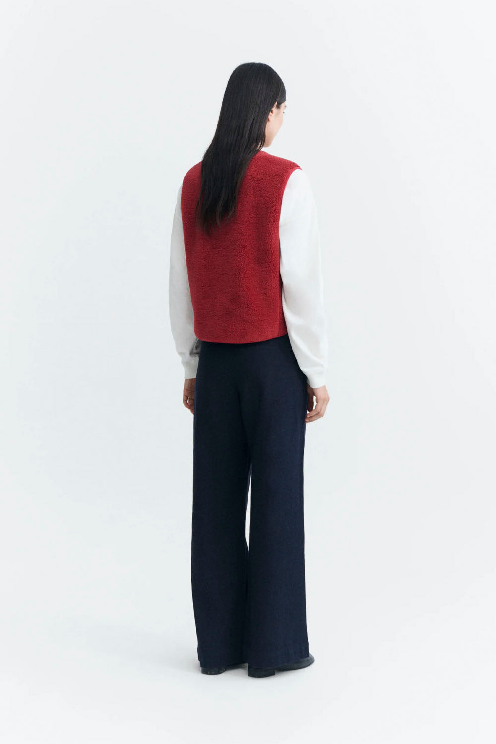 A model viewed from the back wearing a red knit sleeveless vest over a white long-sleeve top and dark navy wide-leg trousers, set against a plain white studio background.