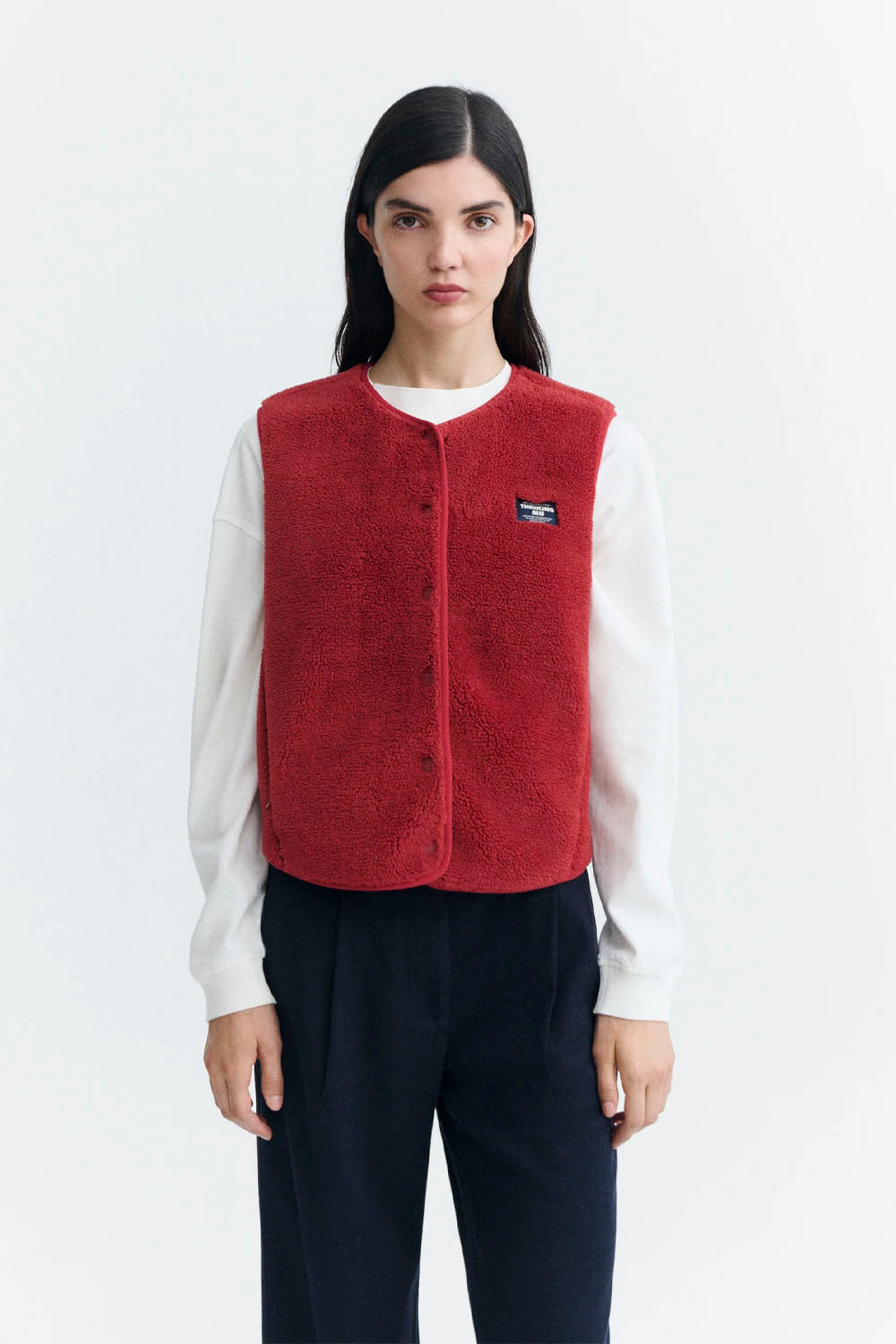 A model wearing a red fleece button-up vest over a white long-sleeve shirt and dark trousers, standing against a light gray studio background.