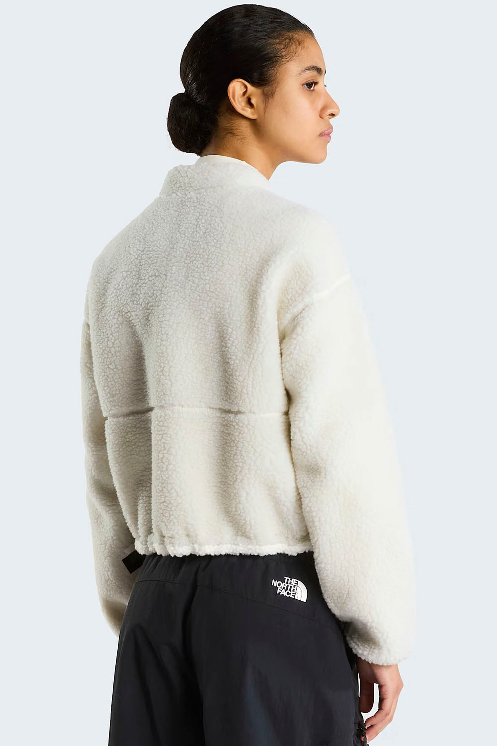 A model shown from the back wearing a cream-colored, textured fleece cropped jacket and black pants with a white The North Face logo.