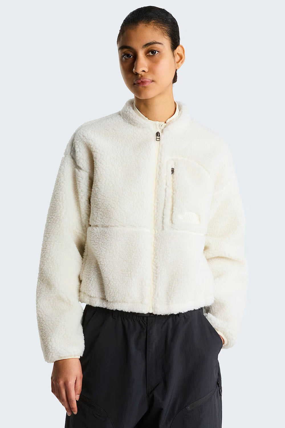 A model wearing a cream-colored sherpa fleece cropped zip-up jacket and dark charcoal pants, photographed against a light gray studio background.