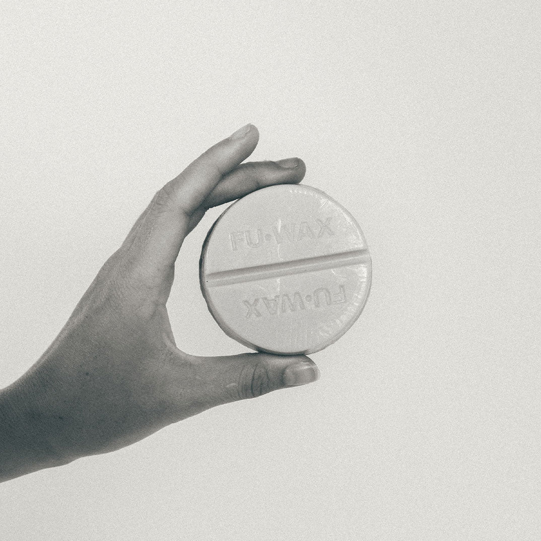 Hand holding a Fu Wax surf wax on a light gray background